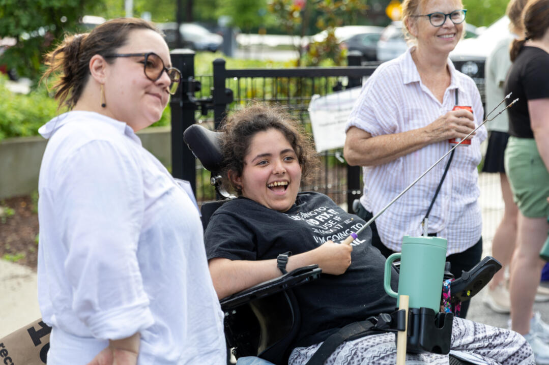 Photo of three people smiling at the camera. The people on the left and right are standing and wearing casual blouses. The person in the middle is sitting in a wheelchair and wearing a black t-shirt. Photo courtesy of Our Stomping Ground