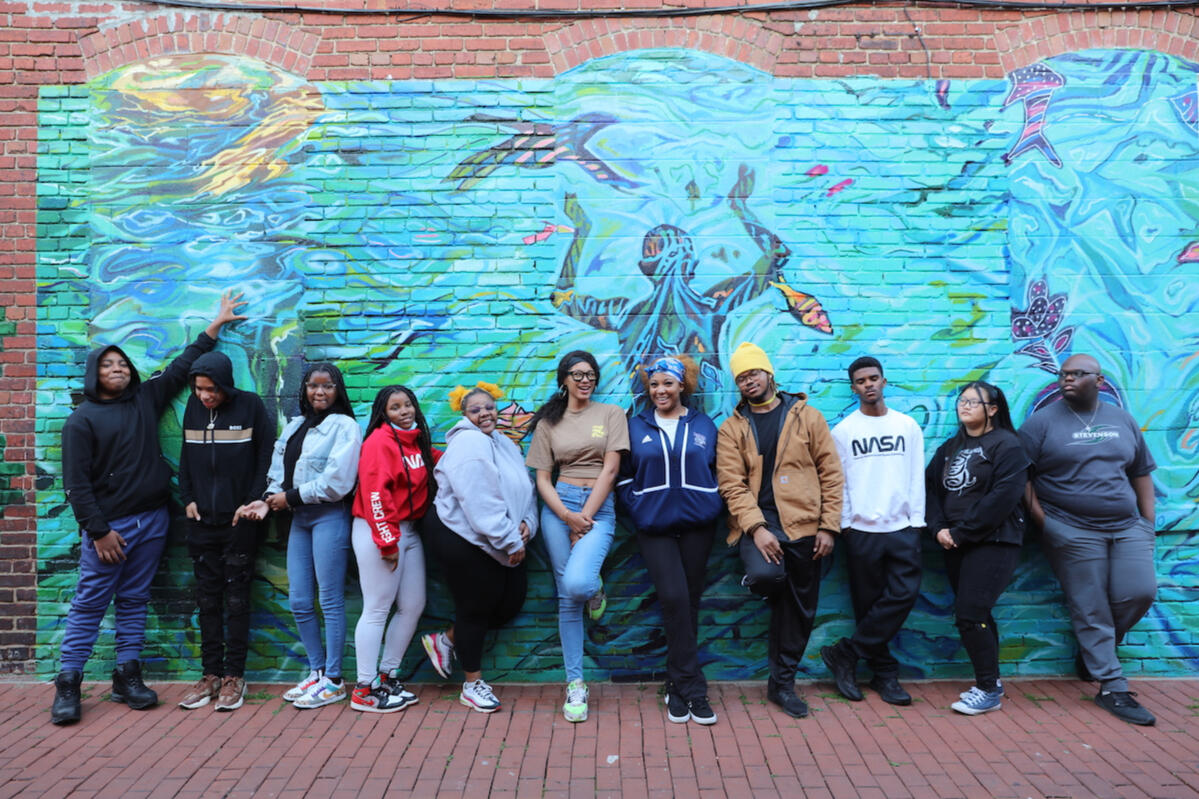 Photo of a group of people posing for the camera in front of a brick mural. The mural is bright blue and shows a person swimming with underwater creatures